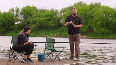 Kafkasyalı rahat bir çift birlikte boş zamanlarını geçiriyorlar, sakin bir gündüz gezisi sırasında nehrin kumlu kıyısında çubuklarla balık avlıyorlar, açık hava hobilerinin ve doğanın tadını çıkarıyorlar.