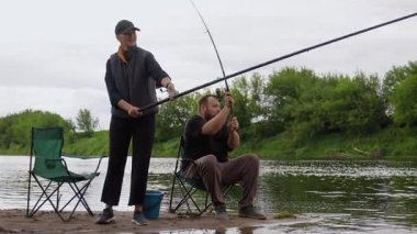 Sakin bir çift bulutlu bir günde nehir kenarında balık tutuyor, olta ve makara tutarak sabırla avlanmayı bekliyor. Yeşil ağaçlar ve bulutlu bir gökyüzü huzur veren doğal bir zemin sağlar.