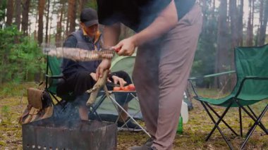 Sakallı bir adam ızgarada marine edilmiş et pişirirken bir kadın çam ormanında kamp alanında yemek hazırlıyor. Doğanın ve yemek pişirmenin tadını çıkaran arkadaşlarla huzurlu bir açık hava aktivitesi.