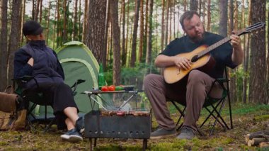 Genç çift, ormanda kamp yaparken kamp ateşinin yanında romantik bir akşam geçiriyorlar. Adam akustik gitar çalıyor ve çadırlarının yanında eşine şarkı söylüyor. Küçük bir piknik masası var.