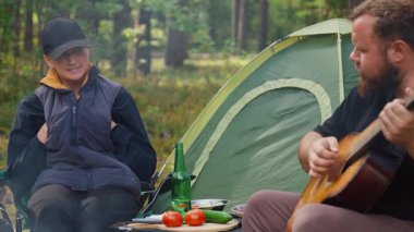 Genç çift, ormanda kamp yaparken kamp ateşinin yanında romantik bir akşam geçiriyorlar. Adam akustik gitar çalıyor ve çadırlarının yanında eşine şarkı söylüyor. Küçük bir piknik masası var.