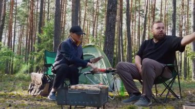 Genç bir çift, güzel bir çam ormanında sandalyelerde otururken pikniğin tadını çıkarıyor. Kadın ekmek dilimliyor ve adam çadırlarının yanındaki masadan sebze ve barbekü alıyor.