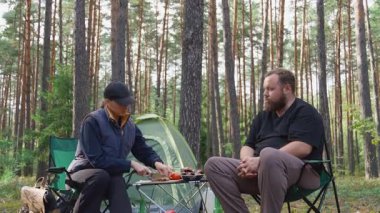Genç bir çift, güzel bir çam ormanında sandalyelerde otururken pikniğin tadını çıkarıyor. Kadın ekmek dilimliyor ve adam çadırlarının yanındaki masadan sebze ve barbekü alıyor.