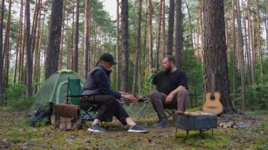Genç bir çift, güzel bir çam ormanında sandalyelerde otururken pikniğin tadını çıkarıyor. Kadın ekmek dilimliyor ve adam çadırlarının yanındaki masadan sebze ve barbekü alıyor.