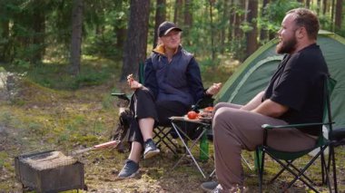 Genç bir çift, güzel bir çam ormanında sandalyelerde otururken pikniğin tadını çıkarıyor. Kadın ekmek dilimliyor ve adam çadırlarının yanındaki masadan sebze ve barbekü alıyor.