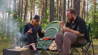Genç bir çift, güzel bir çam ormanında sandalyelerde otururken pikniğin tadını çıkarıyor. Kadın ekmek dilimliyor ve adam çadırlarının yanındaki masadan sebze ve barbekü alıyor.