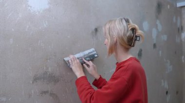Duvarı boyama ve dekorasyona hazırlamak için bağımsız bir ev yenileme projesi sırasında beton yüzeyi dümdüz eden, macun bıçağıyla sıva uygulayan genç sarışın kadın.