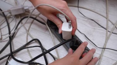 Bir kadının eli, beyaz şarj aletini eski, tozlu ve aşırı yüklü bir güç şeridine takıyor. Etrafı siyah beyaz elektrik kabloları ve adaptörlerle çevrili.