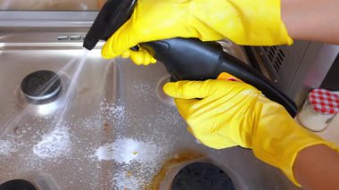 Sarı lastik eldivenli temizlikçi kirli paslanmaz çelik gaz tenceresine deterjan sıkıyor, mutfak yüzeyini temizliyor ve inatçı kiri ve yağı temizliyor.