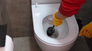 Sarı koruyucu eldivenler giyen temizlikçi, beyaz seramik bir klozeti siyah bir fırçayla iyice temizliyor, modern bir banyoda ev hijyeni ve hijyeni sağlıyor.