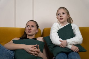 Anne ve kızı yeşil yastıklı sarı bir kanepede birlikte korku filmi izliyorlar, birbirlerine sarılıyorlar ve oturma odasında korku, gerilim ve yakın aile bağlarını gösteriyorlar.