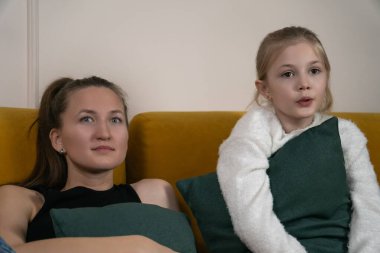 Beyaz anne ve genç kız oturma odalarında sarı bir kanepede oturuyor, birlikte televizyon izliyorlar, rahat, rahat bir bağ kuruyorlar ve evde kaliteli bir ekran zamanı geçiriyorlar.