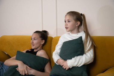 Anne ve kızı sarı bir kanepede, yeşil yastıklar tutarak birlikte dinleniyorlar, oturma odalarında televizyon izliyor ve eğleniyorlar.