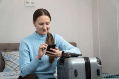 Akıllı telefon kullanan genç bir kadın bavulunun yanında oturuyor, seyahat planlıyor ve online rezervasyon yaptırıyor ya da seyahat bilgilerini kontrol ediyor.