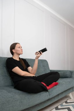 Siyah elbiseli ve kırmızı çoraplı genç bir kadın elinde uzaktan kumandayla bir kanepede oturuyor düşünceli ve sıkılmış bir halde televizyonda ne izleyeceğine karar vermeye çalışıyor.