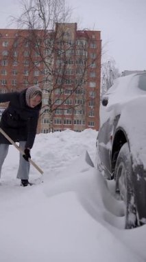 Bir apartmanın arka planında kar arabasının yakınındaki kadın, kalın kar yığınları, bulutlu gökyüzü, katlanabilir ceket ve atkı, sabit ritim, tekerlek temizleme matarası kaldırımı, sert kış atmosferi,