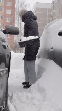 Park halindeki arabanın etrafındaki karları temizleyen, arabaların arasında dar alan, kalın palto ve örgü şapka, sürekli temizlik ve kaldırma, rüzgarda süzülen taneler, paylaşılan dairenin avlusu.