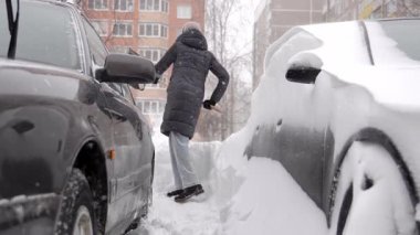 Park halindeki arabanın etrafındaki karları temizleyen, arabaların arasında dar alan, kalın palto ve örgü şapka, sürekli temizlik ve kaldırma, rüzgarda süzülen taneler, paylaşılan dairenin avlusu.