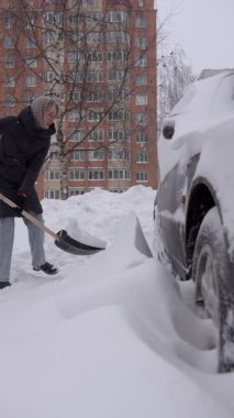 Beyaz kadın park yerindeki araba tekerleği alanını kazıyor, gönüllü enerji odaklı, lastiklerin etrafında ağır sürükleniyor, kış soğuğu ve arka planda uzun tuğla bina, sürekli tekrarlanan kepçeler.