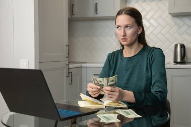 Genç düşünceli bir kadın cam mutfak masasında dizüstü bilgisayarı ve not defteriyle oturmuş bize kalan son dolar banknotlarını sayıyor, finansal sorunları var, borçları düşünüyor ve faturaları ödemek için çabalıyor.