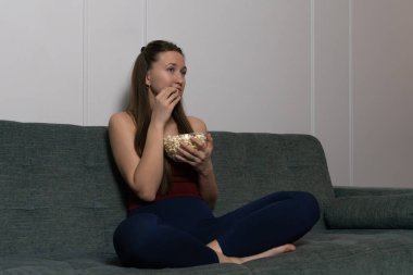 Genç bir kadın rahat bir koltukta oturmuş elinde patlamış mısırla, televizyonda sıkıcı bir film seyrediyor, yüzündeki hayal kırıklığı ve sıkılma ifadesini sergiliyor.