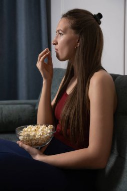 Genç bir kadın kanepede dinleniyor, sıcak bir oturma odasında televizyon izlerken cam bir kaseden patlamış mısırın tadını çıkarıyor, film gecesi için mükemmel bir ortam yaratıyor. Dikey fotoğraf