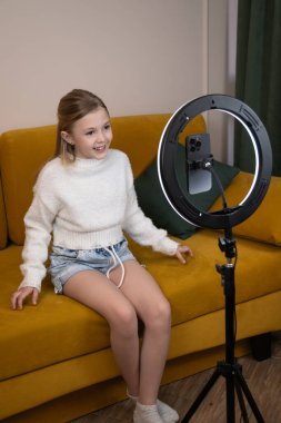 Evdeki bir kanepede oturan genç kız, halka lambasındaki akıllı telefonu kullanarak sosyal medya kanalı için canlı bir video kaydediyor, doğrudan kameraya bakıyor. Dikey fotoğraf