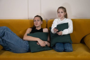 Küçük anne ve kızı rahat bir oturma odasında sarı bir kanepede oturuyor, yastıkları sıkıca tutuyor ve uzaktan kumandayla TV 'ye odaklanıyor.