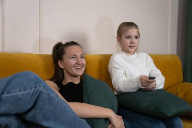 Young woman and little girl relaxing on mustard yellow sofa in cozy living room, smiling and watching TV together during family screen time and bonding moments