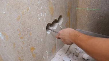 Profesyonel elektrikçi, ev tadilatı sırasında keski ve çekiç kullanarak elektrik prizleri için beton duvarda delikler açıyor ve yeşil lazer düzeyi doğru hizayı gösteriyor.