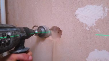 Profesyonel bir işçi, elektrik prizi için beton duvarda delik açmak için güçlü bir çekiç matkabı kullanıyor. Doğru hizalama için yeşil lazer seviyesi rehberliğinde.