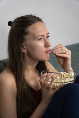 Genç bir kadın kanepede oturur, cam bir kaseden patlamış mısır yer ve büyüleyici bir film izler, ekranda gösterilen olaya kendini tamamen kaptırmıştır. Dikey fotoğraf