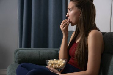 Genç bir kadın oturma odasında kanepede oturmuş cam bir kaseden patlamış mısır yiyor ve televizyonda şüpheli ve korkunç bir film izliyor.