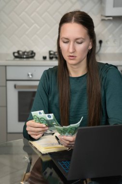 İş kadını ofis masasındaki avro banknotlarını sayarken finans, kazanç hesapları ve finansal planlama için bir dizüstü bilgisayarla bütçesini düzenliyor. Dikey fotoğraf