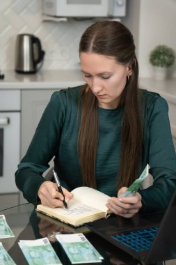 Bilgisayarı kullanarak ve mutfak masasında euro banknotlarını sayarak ev giderlerini hesaplarken, ev bütçesini ve kişisel finansmanı yöneten odaklı kadın defterine notlar yazıyor.