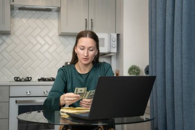 Ev ekonomisini yöneten genç bir kadın. Mutfak masasında dizüstü bilgisayar ve defterle oturup finansal sorumluluk ve ev bütçesini gösterirken bize dolar banknotlarını dikkatlice sayıyor.