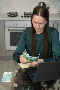 Genç bir kadın ev bütçesini hesaplayarak, modern mutfağındaki avro banknotlarını dikkatlice sayıyor ve online bankacılık ve finans yönetimi için bir dizüstü bilgisayar kullanıyor. Dikey fotoğraf