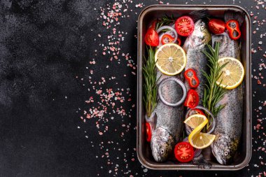 Çiğ alabalık, koyu beton bir masada siyah bir taş kesme tahtasının üzerinde yatar. Yakınlarda: domates, biberiye, limon, biber, soğan, tuz