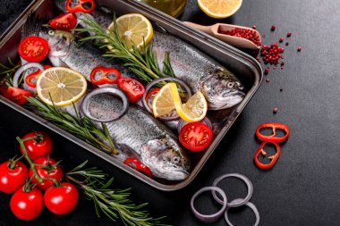 Çiğ alabalık, koyu beton bir masada siyah bir taş kesme tahtasının üzerinde yatar. Yakınlarda: domates, biberiye, limon, biber, soğan, tuz