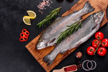 Çiğ alabalık, koyu beton bir masada siyah bir taş kesme tahtasının üzerinde yatar. Yakınlarda: domates, biberiye, limon, biber, soğan, tuz