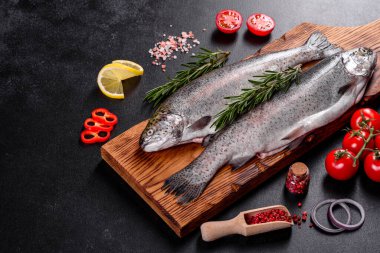 Çiğ alabalık, koyu beton bir masada siyah bir taş kesme tahtasının üzerinde yatar. Yakınlarda: domates, biberiye, limon, biber, soğan, tuz