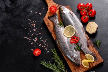 Çiğ alabalık, koyu beton bir masada siyah bir taş kesme tahtasının üzerinde yatar. Yakınlarda: domates, biberiye, limon, biber, soğan, tuz