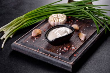 Sarımsak sosu seramik siyah sosta. Koyu beton bir masada ahşap kesme tahtasının üzerinde yeşillik var.