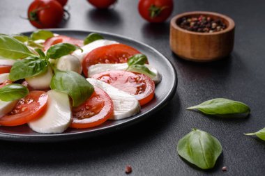 Dilimlenmiş domatesli İtalyan caprese salatası, mozzarella peyniri, fesleğen, zeytinyağı. Eskimiş siyah plaka ile koyu beton arka planda servis ediliyor.