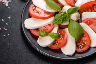 Dilimlenmiş domatesli İtalyan caprese salatası, mozzarella peyniri, fesleğen, zeytinyağı. Eskimiş siyah plaka ile koyu beton arka planda servis ediliyor.