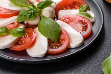 Dilimlenmiş domatesli İtalyan caprese salatası, mozzarella peyniri, fesleğen, zeytinyağı. Eskimiş siyah plaka ile koyu beton arka planda servis ediliyor.