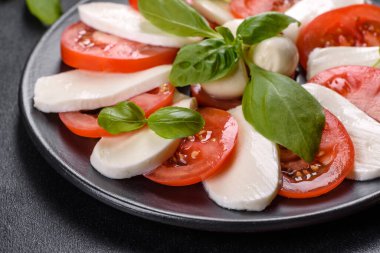 Dilimlenmiş domatesli İtalyan caprese salatası, mozzarella peyniri, fesleğen, zeytinyağı. Eskimiş siyah plaka ile koyu beton arka planda servis ediliyor.