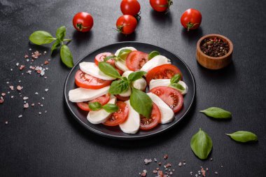 Dilimlenmiş domatesli İtalyan caprese salatası, mozzarella peyniri, fesleğen, zeytinyağı. Eskimiş siyah plaka ile koyu beton arka planda servis ediliyor.