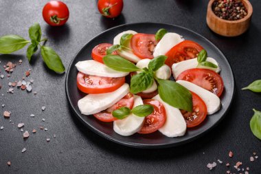 Dilimlenmiş domatesli İtalyan caprese salatası, mozzarella peyniri, fesleğen, zeytinyağı. Eskimiş siyah plaka ile koyu beton arka planda servis ediliyor.