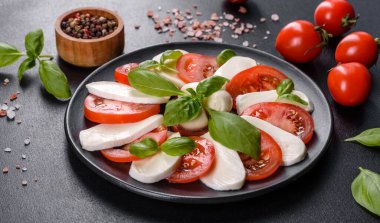 Dilimlenmiş domatesli İtalyan caprese salatası, mozzarella peyniri, fesleğen, zeytinyağı. Eskimiş siyah plaka ile koyu beton arka planda servis ediliyor.